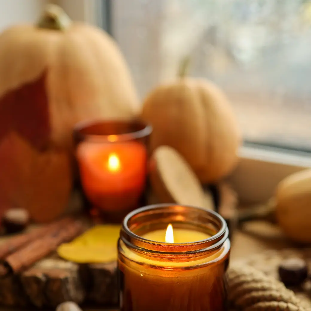 An amber jar candle burning beside pale pumpkins on a windowsill