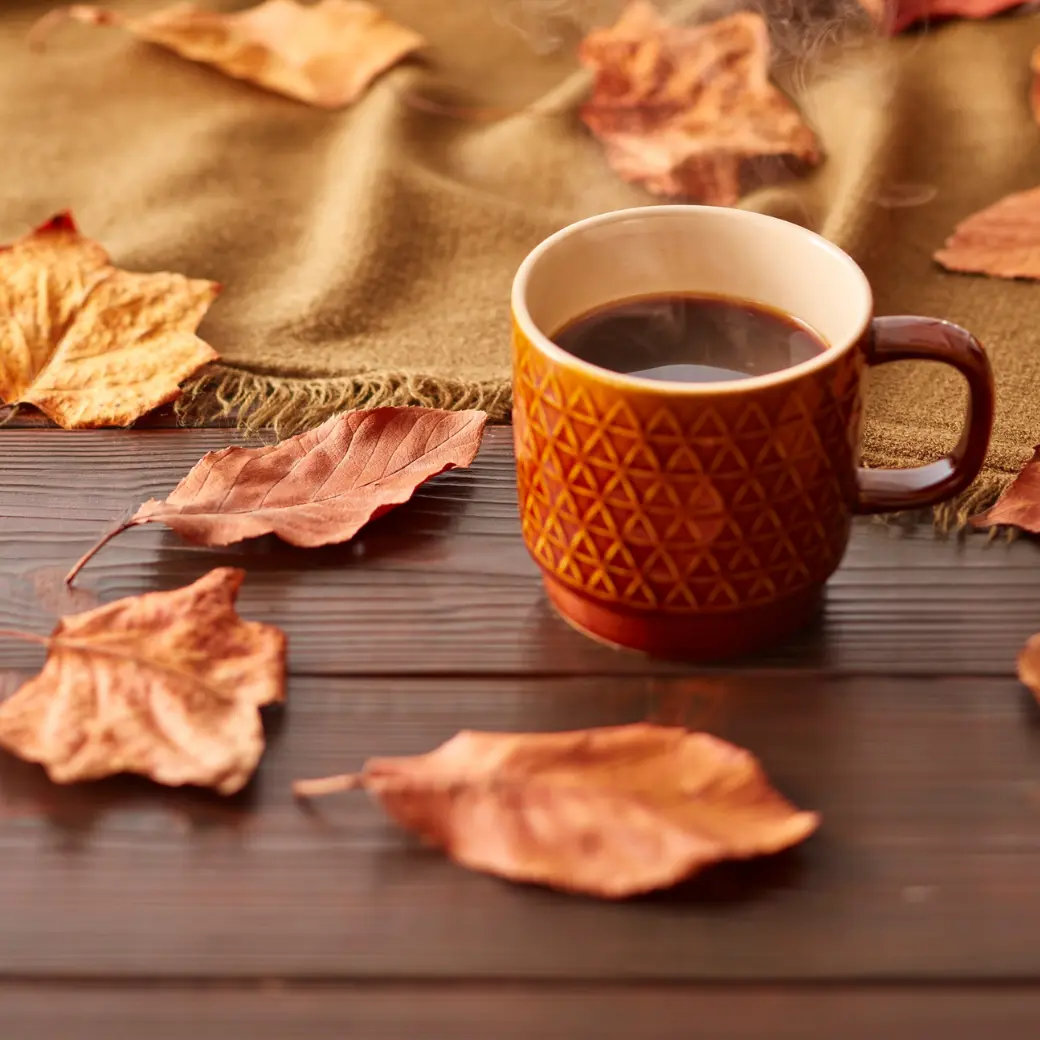 A steaming patterned mug of coffee surrounded by autumn leaves