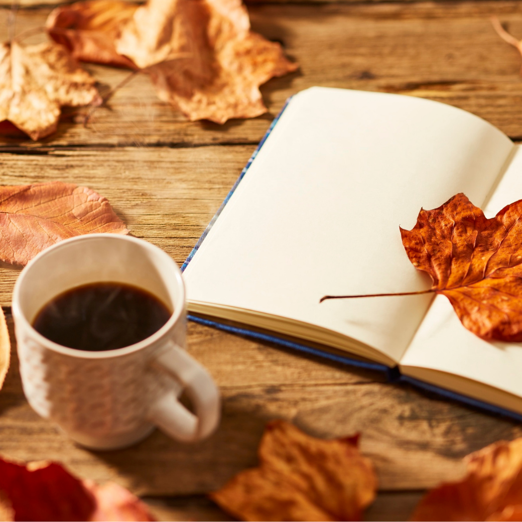 Open journal, mug of coffee, and fallen autumn leaves on a wooden table
