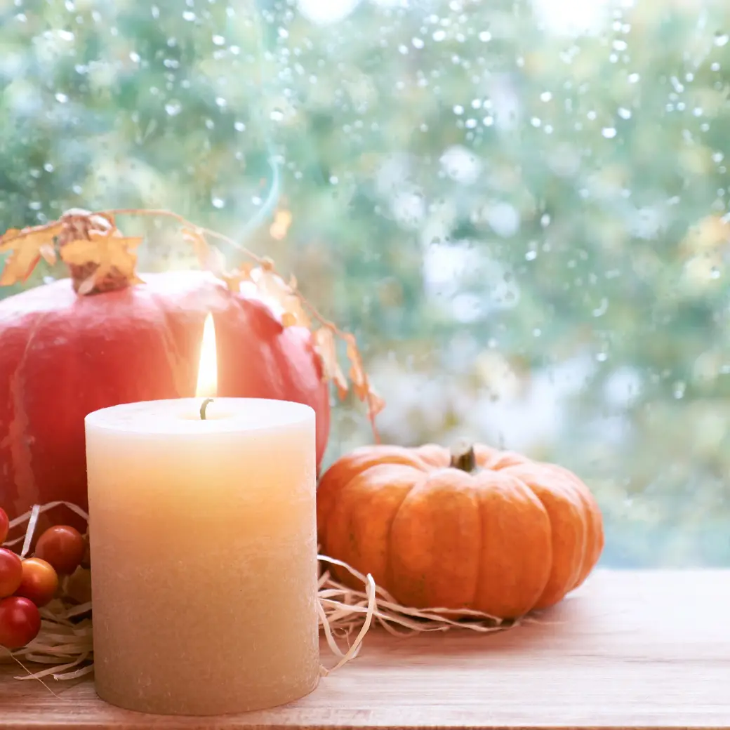 A lit pillar candle beside red and orange pumpkins at a rain-speckled window